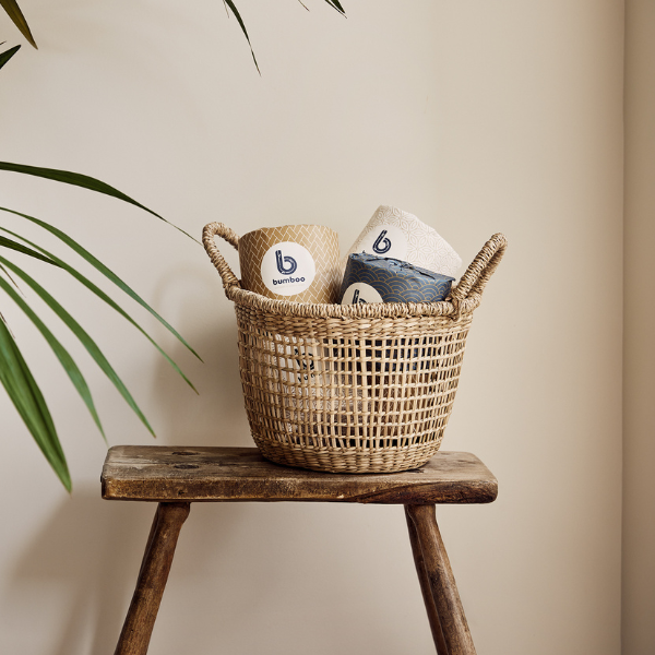 Wrapped bamboo toilet rolls in wicker basket on wooden bench with plant in foreground