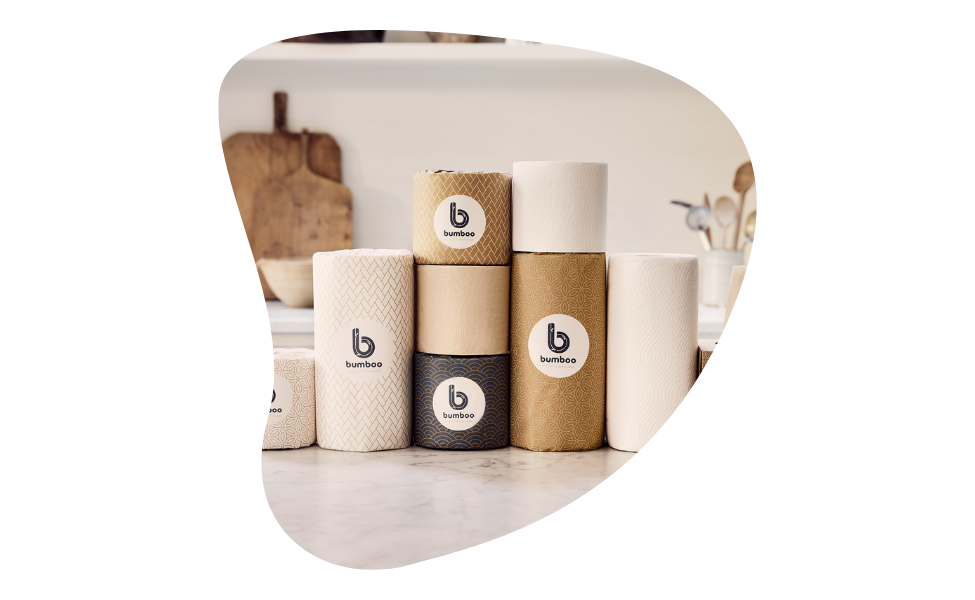 The image shows a kitchen setting with a clean, minimalist design. In the foreground, there is a variety of paper products, including toilet paper rolls, paper towels, and tissues, all from the brand "bumboo." The products are arranged neatly on a marble countertop. Each product is wrapped in eco-friendly packaging featuring natural colors like beige, brown, and black, with the brand logo prominently displayed.  In the background, the kitchen has a white theme with wooden elements, including cutting boards 